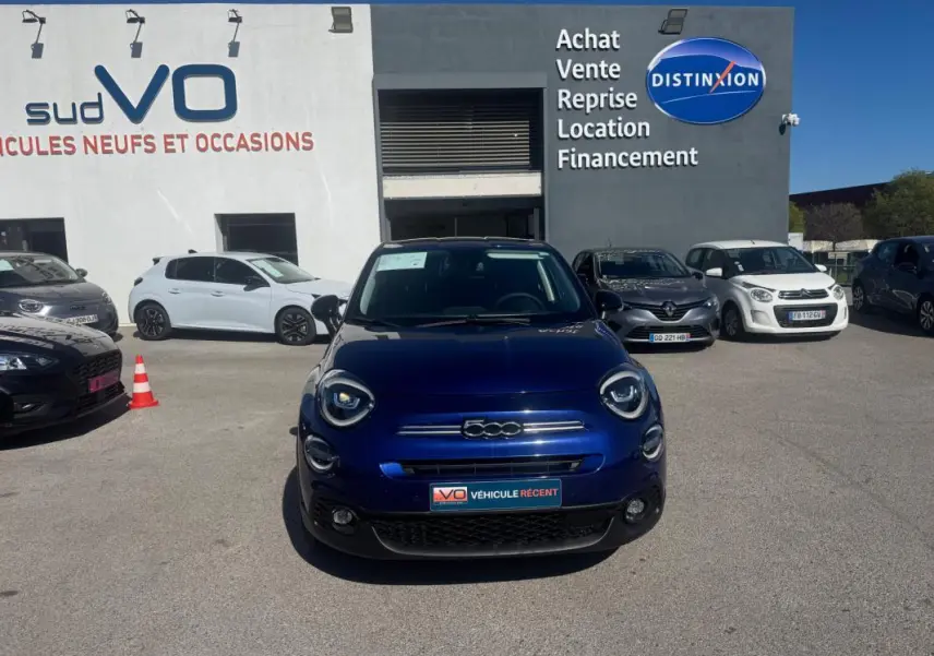 Vue avant d'une FIAT 500X bleu profond MY23, avec phares ronds et calandre chromée, en extérieur sur parking.