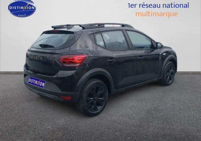Vue 3/4 arrière droite d'une Dacia Sandero noire avec barres de toit et jantes alu noires sur fond neutre.