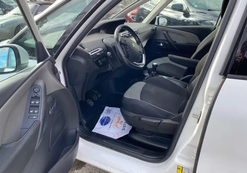 Intérieur côté conducteur d'un Citroën Grand C4 Picasso blanc, montrant le volant cuir, levier de vitesse manuel et sièges tissu gris.
