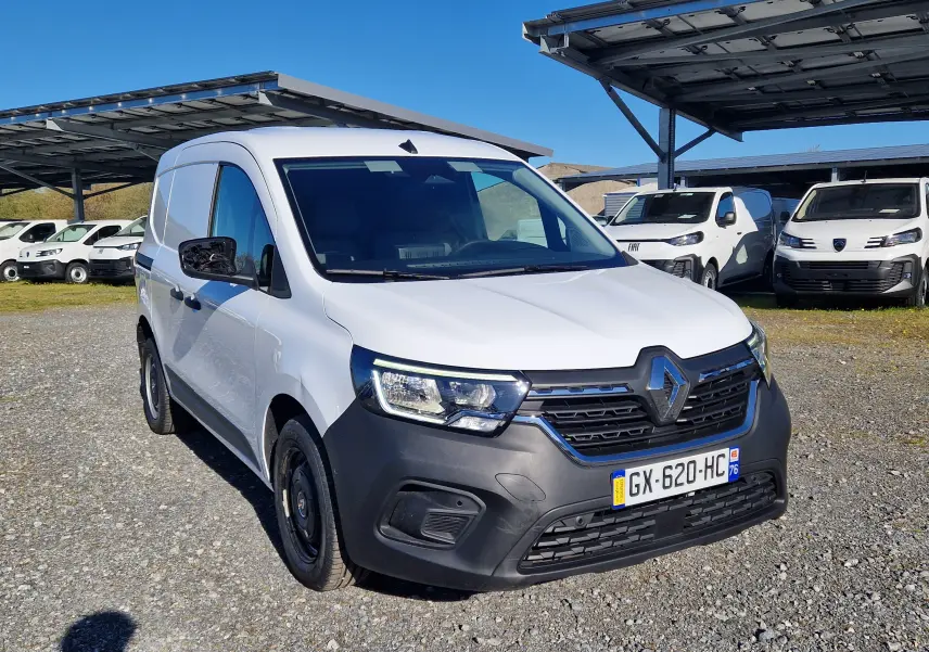 Vue 3/4 avant droite d’un Renault Kangoo blanc 2024 avec pare-chocs noir et calandre chromée, stationné sur un sol gravillonné.