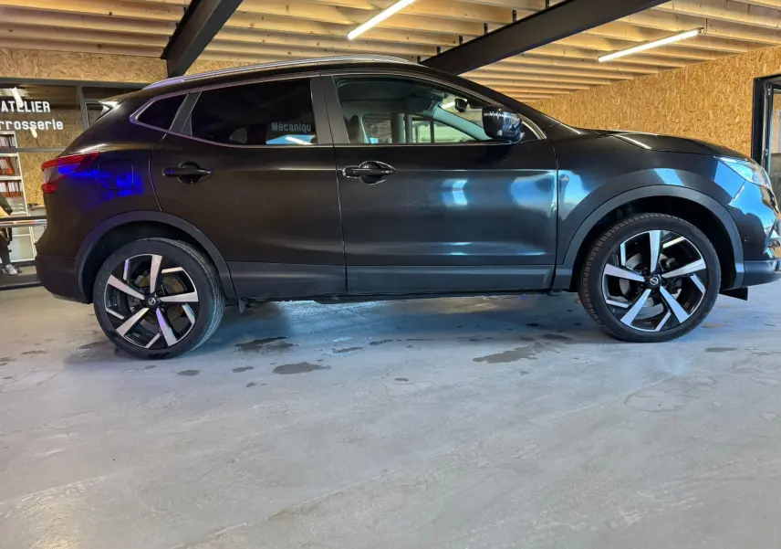 Vue de profil côté gauche d'un Nissan Qashqai noir 2017 avec jantes alliage et barres de toit en intérieur d'atelier.