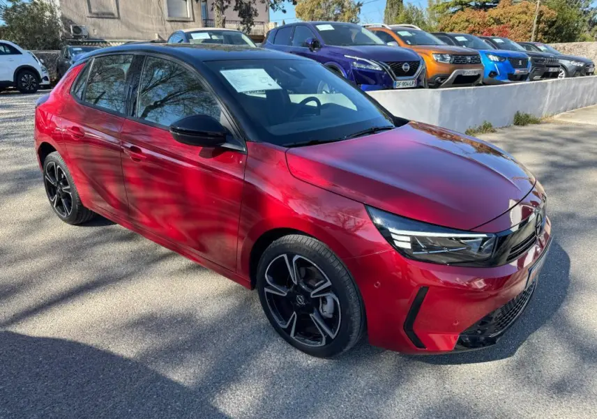 Opel Corsa rouge vue 3/4 avant droit, jantes noires et toit noir, stationnée en extérieur au soleil.