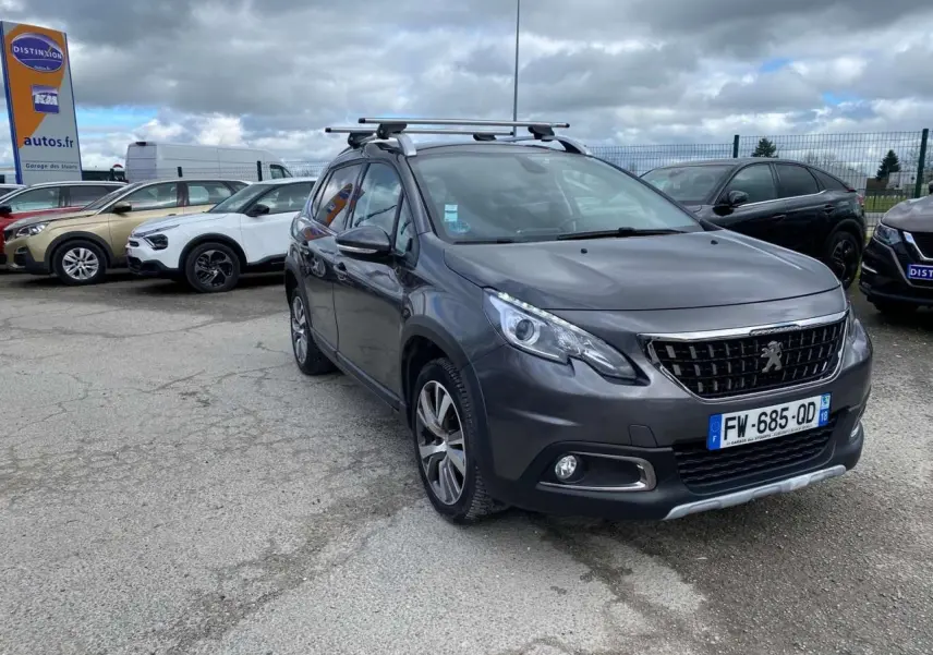 Peugeot 2008 gris platinium vue 3/4 avant droit avec barres de toit aluminium sur parking extérieur.