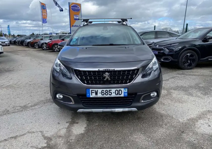 Vue frontale d'un Peugeot 2008 gris platinium avec barres de toit aluminium sur un parking extérieur.