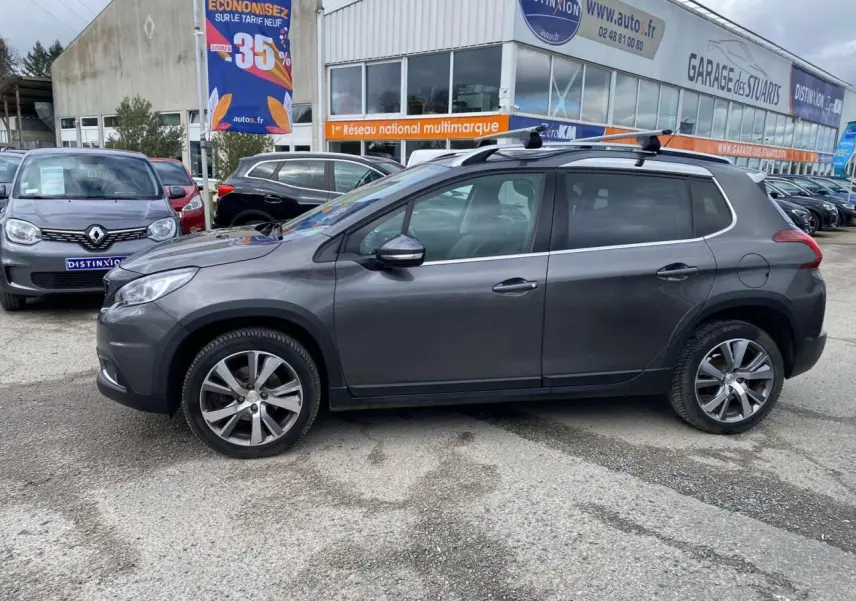 Profil côté gauche du Peugeot 2008 gris platinium 2019 avec barres de toit aluminium sur parking extérieur.