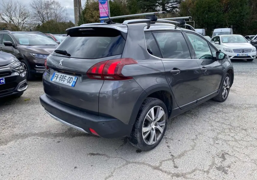 Peugeot 2008 gris platinium vue 3/4 arrière droit avec barres de toit aluminium sur parking.