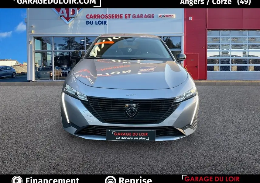 Vue avant d'une Peugeot 308 2024 gris Artense avec calandre noire et feux LED, stationnée devant un garage.