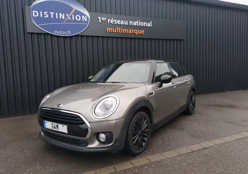 MINI Clubman F54 Silver Metal en 3/4 avant droit, toit noir et jantes foncées sur parking devant un mur noir.