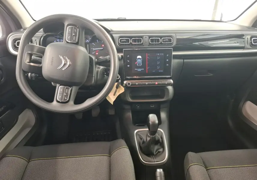 Intérieur de Citroën C3 2024 vu de face, tableau de bord noir brillant avec volant multifonctions et sellerie tissu gris à surpiqûres jaunes.