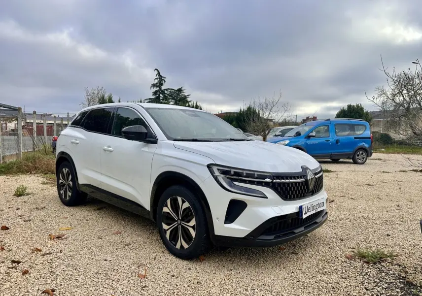 Renault Austral blanc en 3/4 avant droit, avec calandre noire et jantes modernes sur un sol gravillonné.