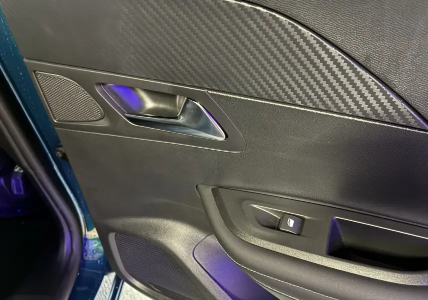 Détail de la porte intérieure côté gauche d’un Peugeot 2008 bleu 2025, avec poignée métal et insert effet carbone.