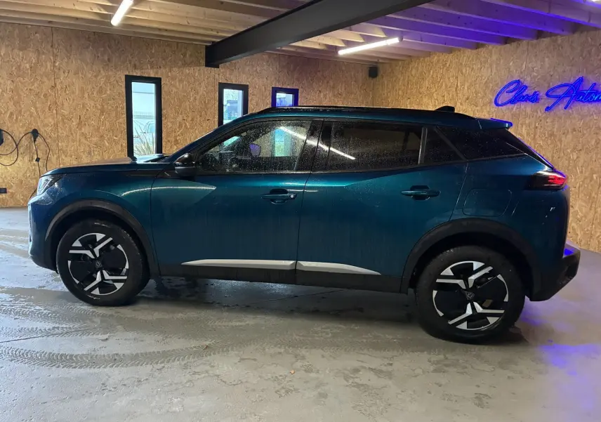 Profil latéral droit d'un Peugeot 2008 bleu hybride 2025, avec jantes noires et toit noir contrastant en intérieur d'atelier.