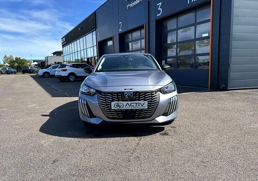Peugeot 208 1.2 100 Allure gris Artense vue de face, avec calandre noire distinctive et phares LED allumés.
