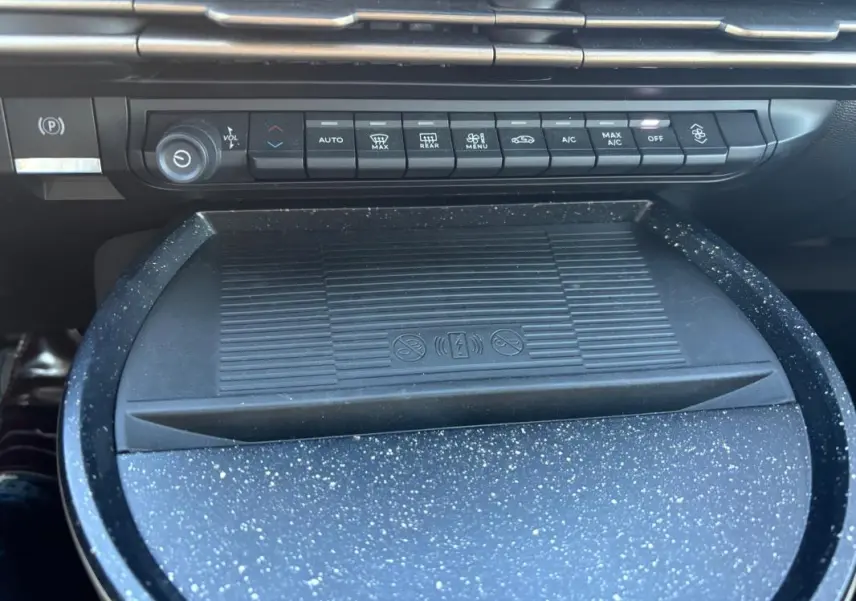 Vue rapprochée de la console centrale de la Lancia Ypsilon 2025 avec commandes climatisation et surface de recharge sans fil noire à motifs gris.