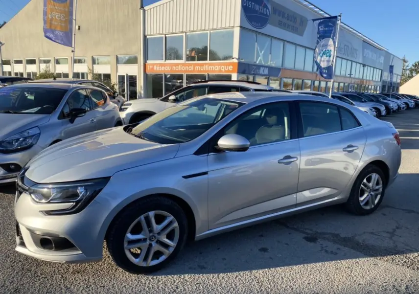 Renault Mégane Sedan gris platine vue de profil côté droit, stationnée devant un concessionnaire multimarque.