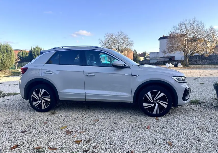 Volkswagen T-Roc gris vue de profil côté gauche sur un sol gravillonné avec arrière-plan rural.