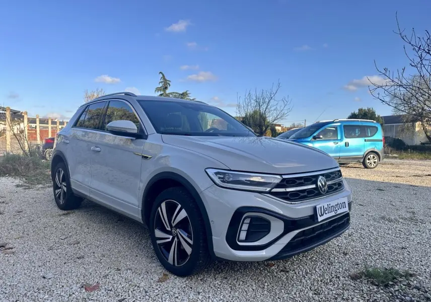 Volkswagen T-Roc gris en 3/4 avant droit, avec jantes R-Line et calandre noire distinctive.
