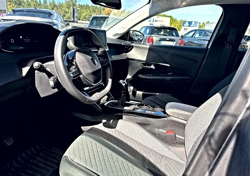 Vue intérieure côté conducteur de la Peugeot 208 1.2 100 Allure 2025 avec sellerie tissu gris et volant compact multifonction.