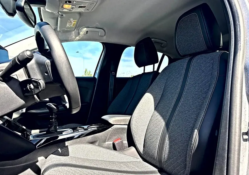 Vue intérieure côté conducteur de la Peugeot 208 1.2 100 allure 2025, sièges tissu gris et volant noir.