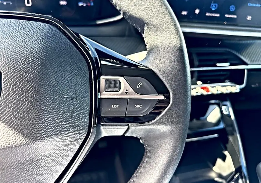 Gros plan sur le volant cuir noir de la Peugeot 208 1.2 100 Allure, avec commandes audio et tableau de bord numérique.