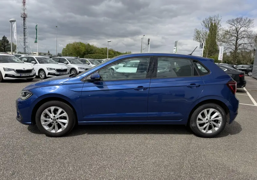 Vue de profil côté gauche d'une Volkswagen Polo bleue garée sur un parking extérieur sous un ciel nuageux.