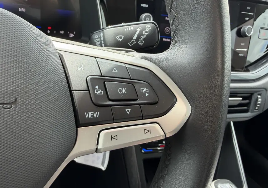 Gros plan sur les commandes multifonctions en cuir noir du volant de la Volkswagen Polo 2025, avec écran tactile en arrière-plan.