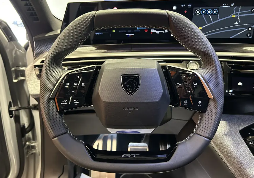 Vue rapprochée du volant multifonction noir perforé du Peugeot 3008 III Hybrid GT blanc Okenite avec tableau de bord numérique.