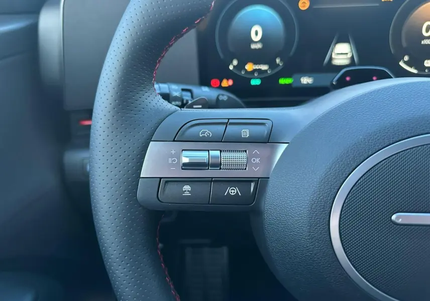 Gros plan sur les commandes gauche du volant cuir perforé avec surpiqûres rouges du Hyundai KONA II noir Abyss.