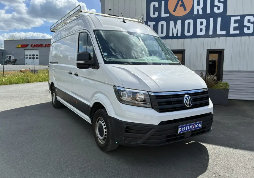 Vue 3/4 avant d’un Volkswagen Crafter blanc avec galerie de toit, stationné devant un bâtiment commercial.