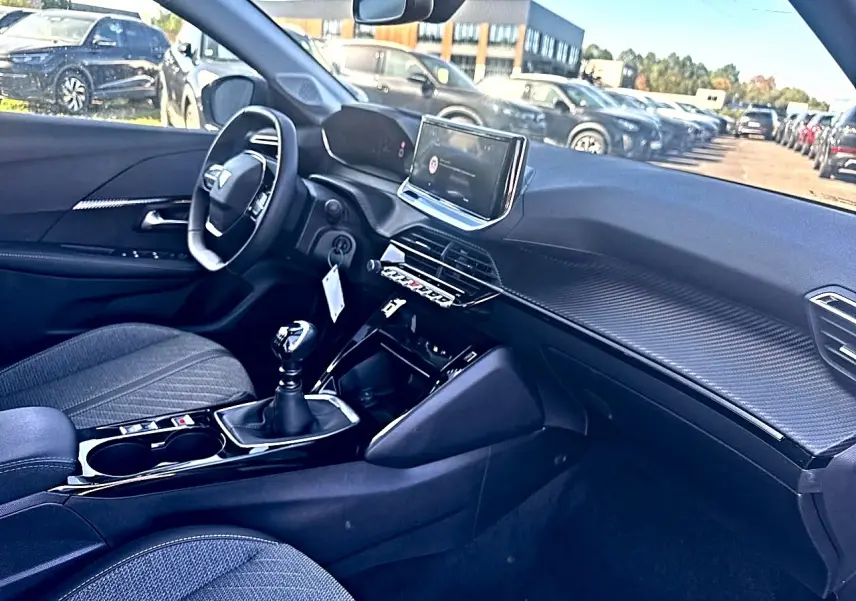 Vue intérieure côté conducteur de la Peugeot 208 noire, avec tableau de bord moderne et boîte manuelle visible.