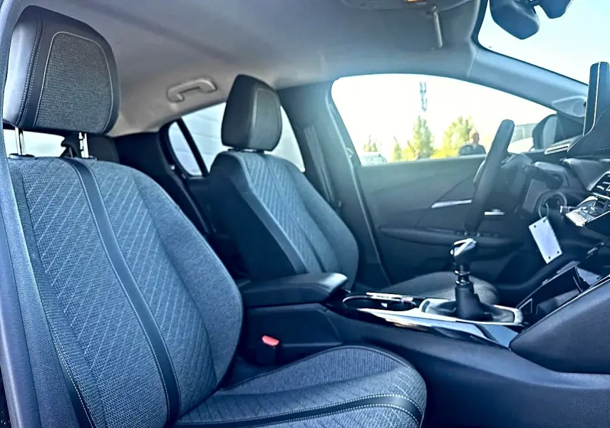 Vue intérieure côté gauche de la Peugeot 208 1.2 100 allure 2025, sièges en tissu gris et boîte manuelle visible.