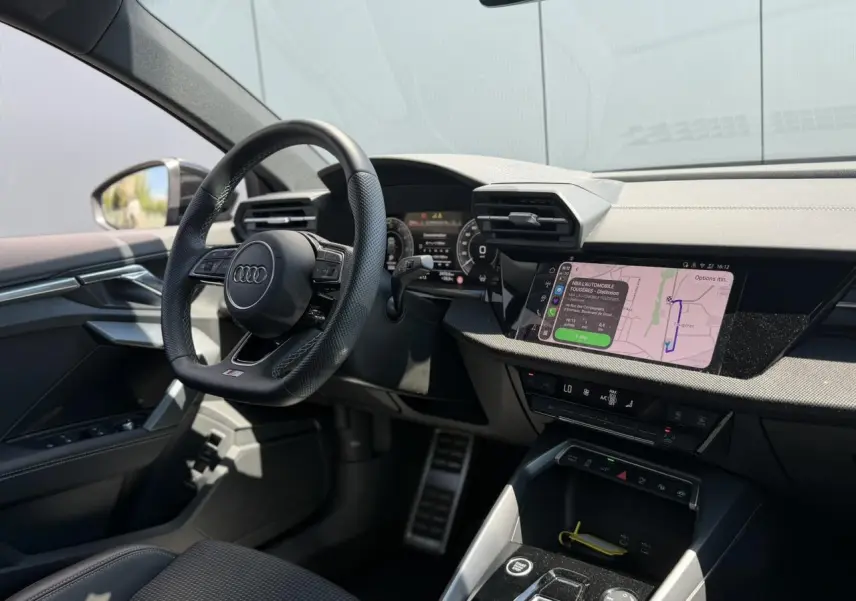 Vue intérieure côté conducteur de l'Audi A3 Sportback 2025, volant cuir perforé et écran tactile de navigation allumé.