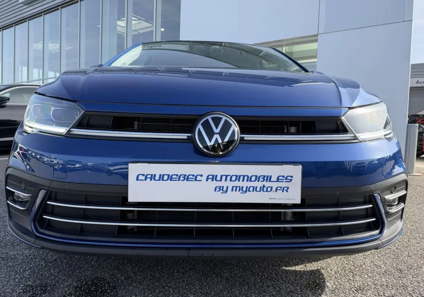 Vue frontale rapprochée d'une Volkswagen Polo 2025 bleu récif métallisé avec logo VW centré et phares LED allumés.