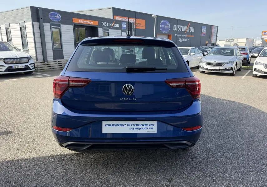 Vue arrière d'une Volkswagen Polo bleue métallisée stationnée devant un concessionnaire avec plusieurs autres voitures visibles.