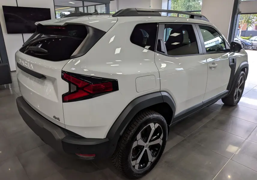 Vue 3/4 arrière droite du Dacia Duster Hybrid-G blanc Glacier, avec jantes bi-ton et protections noires contrastantes.