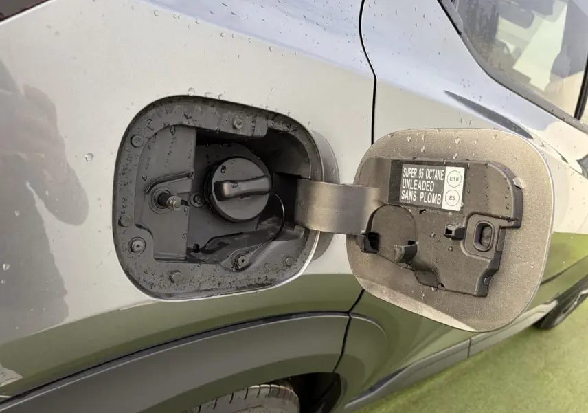 Gros plan sur la trappe à carburant ouverte côté gauche d’un Nissan Juke II noir métal avec pluie visible.
