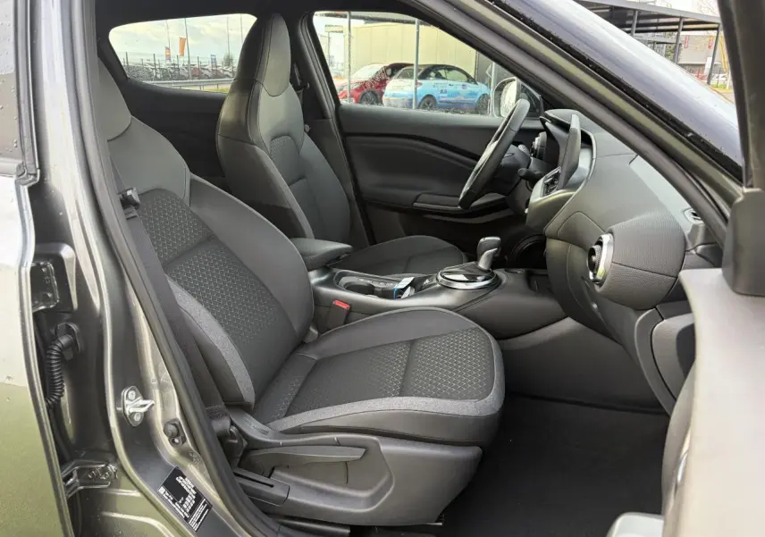 Vue intérieure côté conducteur du Nissan Juke II 1.6 Hybrid noir métal, avec sièges tissu et console centrale moderne.