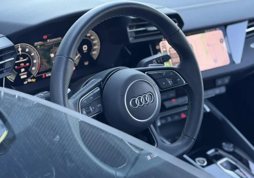 Vue rapprochée du volant cuir perforé noir avec logo Audi et tableau de bord numérique de l’Audi A3 Sportback 2025.