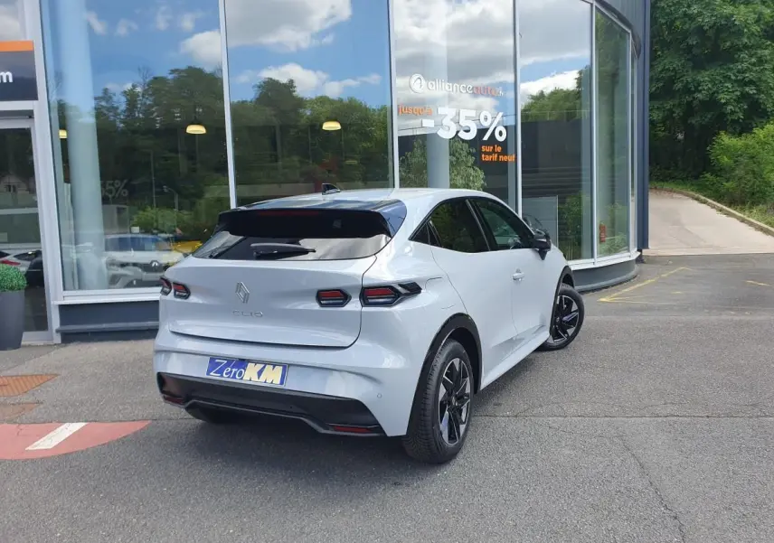Vue 3/4 arrière droite d'une Renault Clio VI gris Rafale avec jantes alliage noir devant un bâtiment vitré.