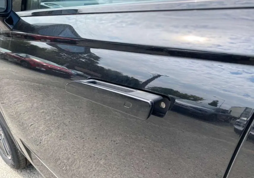Poignée de porte rétractable côté gauche du JAECOO J7 2026 en noir Carbon Cristal, reflet du ciel et environnement urbain.