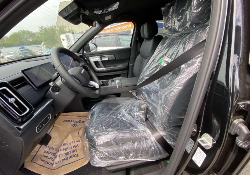 Vue intérieure côté conducteur du JAECOO J7 1.5 PHEV Selective 2026 en noir Carbon Cristal, sièges protégés par film plastique.
