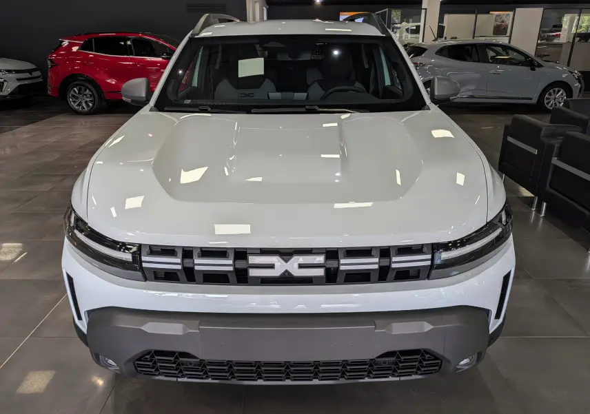 Vue frontale d'un Dacia Duster Hybrid-G blanc glacier avec calandre noire et pare-chocs gris en showroom.