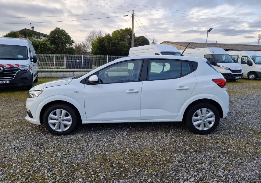 Profil côté gauche d'une Dacia Sandero blanche 2022, version 1.0 ECO-G 100 Confort, sur un sol gravillonné en extérieur.