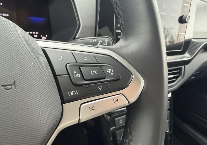 Gros plan sur le volant multifonction cuir noir du Volkswagen T-Cross 2025, avec commandes intégrées et écran tactile en arrière-plan.