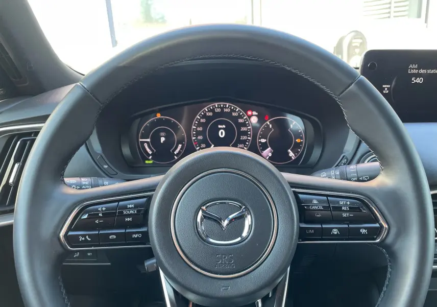 Vue rapprochée du volant cuir noir du Mazda CX-80 2024 avec commandes intégrées et tableau de bord numérique.