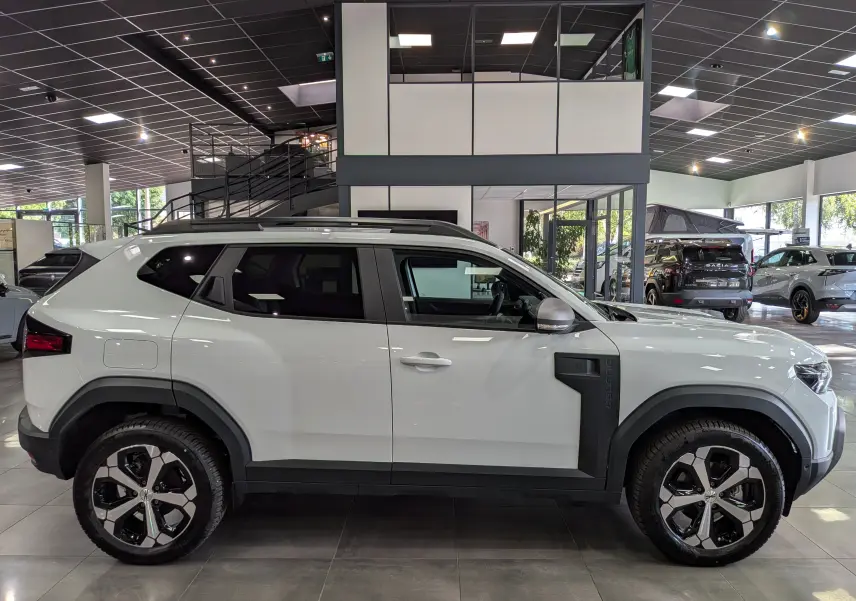 Vue de profil côté gauche d'un Dacia Duster Hybrid-G blanc Glacier en showroom, avec jantes bi-ton et protections noires sur les ailes.