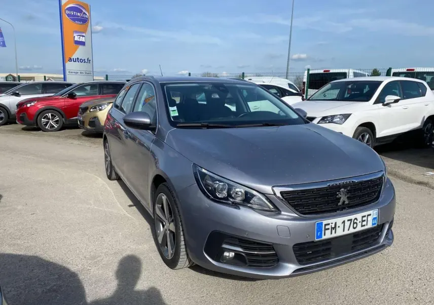 Peugeot 308 SW gris Artense métal vue 3/4 avant droit sur parking avec calandre noire et phares LED.