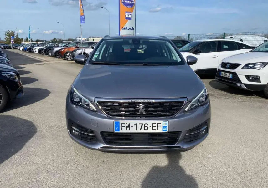 Vue frontale d'une Peugeot 308 SW gris Artense métallisé stationnée sur un parking extérieur avec plusieurs autres véhicules.