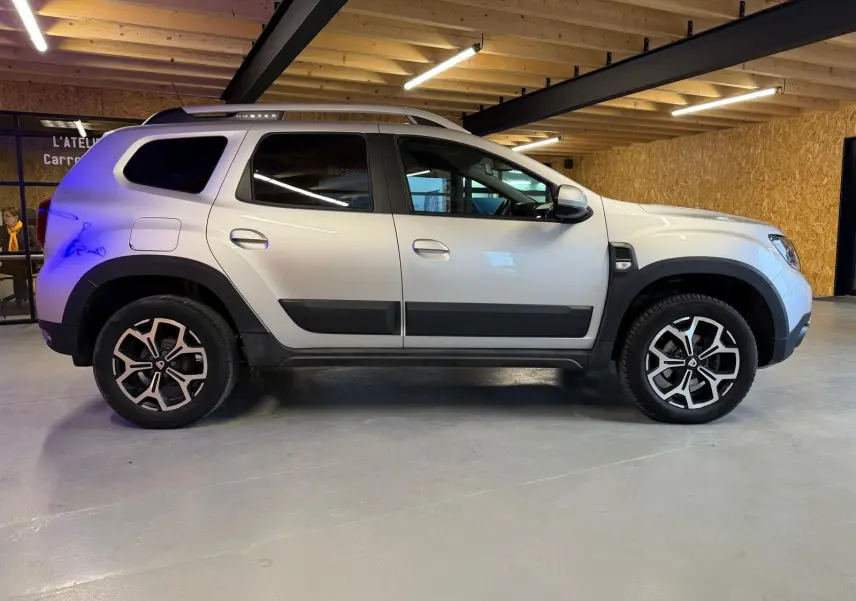 Profil côté gauche d'un Dacia Duster gris 2019 avec jantes alliage 17'' Maldives et barres de toit visibles.
