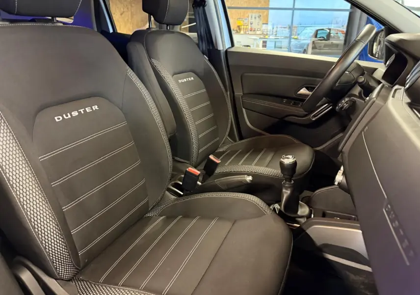 Intérieur avant du Dacia Duster 2019, sièges noirs "Duster" avec surpiqûres, volant et levier de vitesse visibles côté droit.
